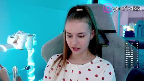 Video of a young Caucasian woman with straight brown hair in a ponytail, wearing a white shirt with red strawberries, sitting in a grey gaming chair, with a blue background and gaming monitor in the background.