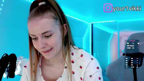 Video of a young woman with fair skin and straight blonde hair tied in a ponytail, wearing a white shirt with red cherry patterns, smiling while recording on a smartphone in a brightly lit room with blue LED lights.