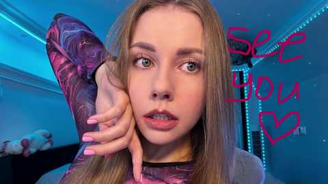 Video of a fair-skinned, blonde-haired young woman with green eyes, wearing a long-sleeved, patterned top, with \"SEE YOU\" in pink heart-shaped text on a blue background.