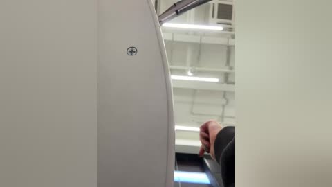 Video of an airplane cabin aisle, featuring a bald man in a black shirt and dark pants walking towards the camera, with white ceiling lights and beige walls in the background.