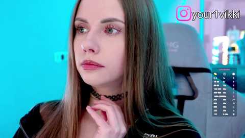 Video of a young woman with long brown hair, wearing a black choker, seated in a gaming chair. Background features a turquoise light and a partially visible PC monitor with gaming controls.
