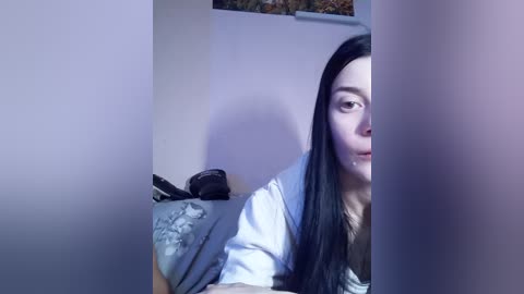 A video of a young woman with long black hair, partially obscured by a vertical object, lying on a bed with gray bedding and a black jacket.