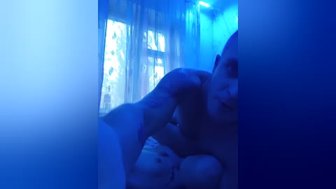 Video of a naked man with a muscular physique, partially obscured by a blue curtain, with a window in the background.