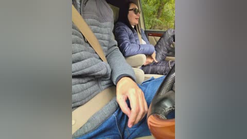 Video of a woman in a dark blue puffer jacket and jeans, sitting in a car seat next to a man in a gray puffer jacket, with a blurred green forest background.