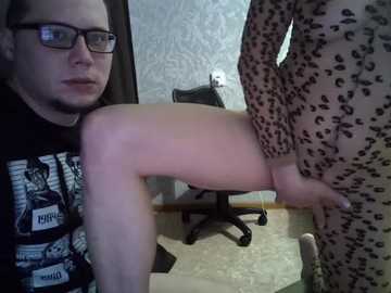 Video of a man in glasses and a graphic t-shirt sitting on a chair, with a woman in leopard print pajamas standing in front of him, both indoors.