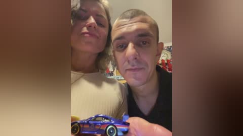 Video of a bald, young man and a woman with curly hair, both holding a blue toy car, set against a blurred background.