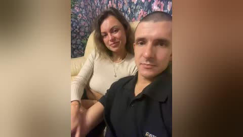 Video of a Caucasian couple on a beige sofa, woman with shoulder-length brown hair, light skin, wearing a cream top, and man with a shaved head, light skin, wearing a black polo shirt. Floral wallpaper in the background.