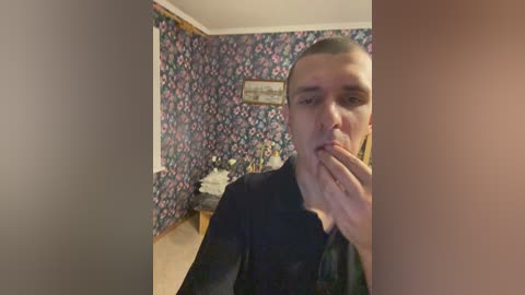 Video of a young man with short hair, wearing a black shirt, standing in a room with floral-patterned wallpaper, wooden floor, and a white framed picture on the wall.
