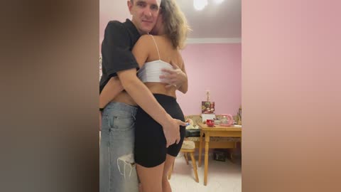 A video of a man and woman in a cozy, pink-painted room, embracing closely; he wears a black shirt and ripped jeans, while she sports a white crop top and black shorts.