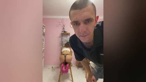 Video of a young, bald man in a black shirt, leaning over a woman in pink lingerie on a wooden chair, in a pink-painted room.