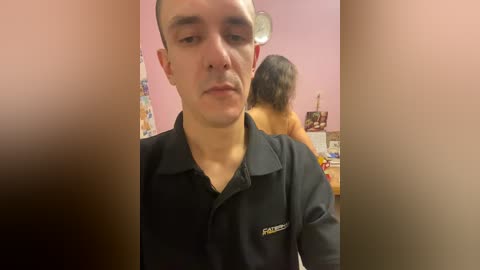 A video of a young man with short hair wearing a black polo shirt, standing in front of a woman with long hair in a pink room.