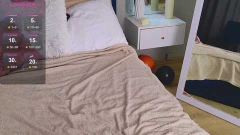 A video of a cozy bedroom with a beige blanket, white nightstand, and a mirror reflecting a black garment.