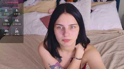 A video of a young woman with straight black hair and fair skin, wearing a red top, sitting on a bed with beige sheets. The background shows a messy room with pillows and a TV displaying a live stream.