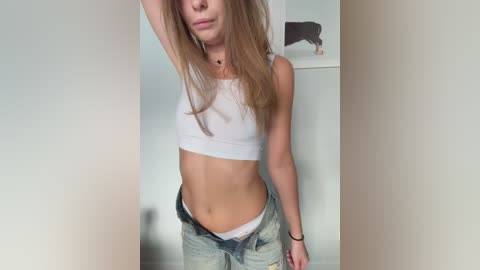 Video of a slim, young woman with long, straight, blonde hair, wearing a white crop top and low-rise, distressed blue jeans, standing in a minimalist room with a white wall and a framed picture of a bull.