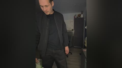 Video of a man in a dark room, wearing a black jacket over a black shirt, holding a glass bottle, looking down. Background includes a kitchen with appliances and cabinets.