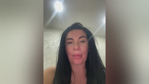 A video of a woman with long black hair, fair skin, and full lips, standing indoors with a bright light source in the background, creating a halo effect.