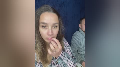 Video of a young woman with straight, light brown hair and light skin, wearing a plaid shirt, biting her finger, with a blurred background.