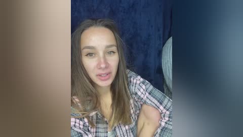 A video of a young woman with long brown hair, wearing a plaid shirt, seated against a dark blue background. She has a light complexion and is smiling slightly.