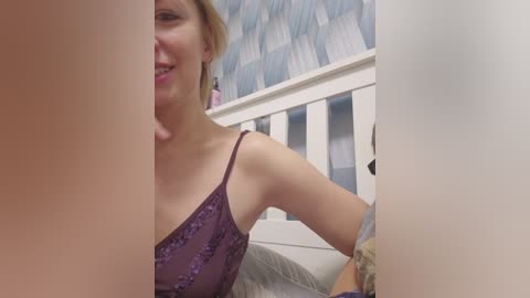 A video of a smiling, light-skinned woman with shoulder-length blonde hair, wearing a purple lace camisole, sitting on a bed with a white headboard and a blue patterned wallpaper.