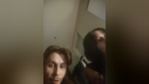 A blurry video of a woman with dark hair, possibly wearing a dark jacket, standing in a dimly lit hallway with beige walls and a ceiling light fixture.