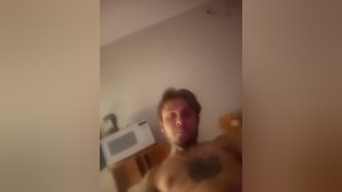 A blurry video of a shirtless man with a beard, standing indoors, near a microwave and kitchen counter. The image is dimly lit, with a warm, orange hue.