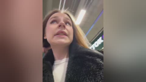 Video of a young woman with fair skin, long blonde hair, and a white shirt, wearing a black fur coat, looking upward with a surprised expression. Background includes a blurred indoor setting with fluorescent lights and indistinct people.