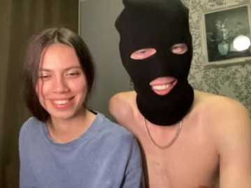 Video of a smiling woman with shoulder-length dark hair in a blue shirt, standing beside a shirtless man wearing a black ski mask.