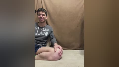 Video of a young, light-skinned man with short dark hair, wearing a grey sweatshirt with \"East Coast JAW\" and shorts, sitting cross-legged on a beige carpet, against a tan backdrop.
