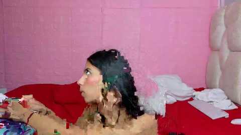 A blurry video of a young woman with dark hair, lying on a bed with a red sheet, surrounded by white clothing, against a pink brick wall.