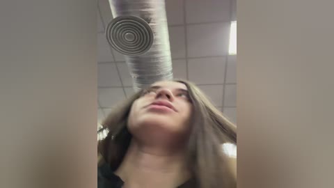 A video of a woman with long brown hair and fair skin, standing under a suspended air duct in a ceiling with fluorescent lights. She looks up with a neutral expression.