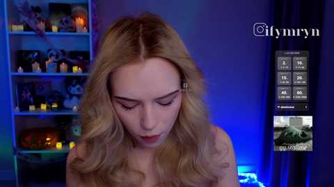 Video of a blonde woman with long hair, wearing a sleeveless top, standing indoors against a blue-lit background with candles, shelves, and a weather app on screen.