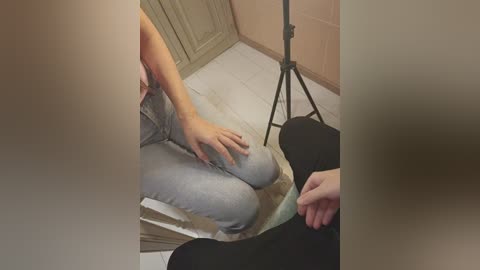 Video of a person sitting on a white tiled floor, wearing light grey sweatpants and a black top. A tripod-mounted camera is set up nearby.
