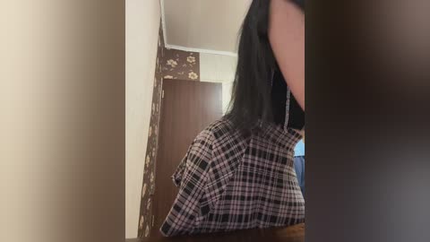 Video of a person with long black hair, wearing a plaid shirt, standing in a dimly lit room with floral wallpaper and a wooden door in the background.