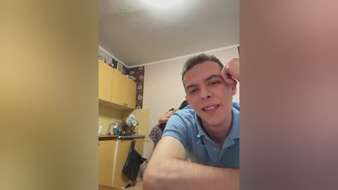 A video of a young man with short hair, smiling, wearing a blue polo shirt, in a small, cluttered room with yellow cabinets, a woman in the background, and a white ceiling light.