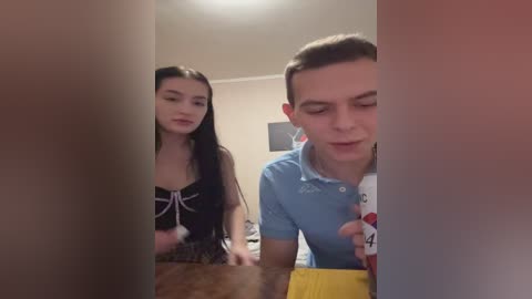 Video of a young woman with long black hair in a black dress and a young man with short brown hair in a light blue polo shirt, both looking down at a dog in a room with beige walls.