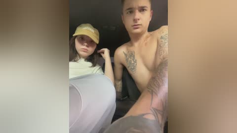 Video of a young woman with long hair in a yellow cap and a shirt, sitting next to a shirtless tattooed man with a stern expression, both in a car.