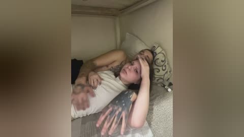 A video captures a young woman with fair skin, long dark hair, and a sleeve tattoo lying on a bed, holding her head in distress. The room is dimly lit, with beige walls and patterned pillows.