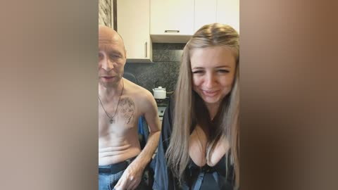 Video of a bald, muscular man with a tattooed chest, wearing blue jeans, and a young woman with long blonde hair, wearing a low-cut black top, standing in a modern kitchen.