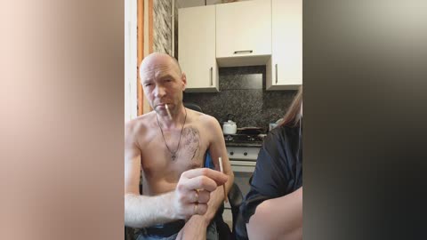 Video of a shirtless, bald man with a tattoo on his chest, sitting in a kitchen with white cabinets, a black granite countertop, and a woman partially visible in the background.