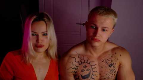 Video of two people: a blonde woman with red lipstick in a low-cut red dress, and a shirtless man with a large sun tattoo on his chest, standing indoors under dim lighting.