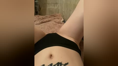 A video of a person lying on a beige blanket, wearing black underwear, with a tattoo of a mountain range visible on their lower abdomen. The background includes a cardboard box.