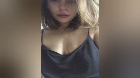 Video of a young woman with shoulder-length brown hair, wearing a black spaghetti-strap tank top, revealing cleavage. She has a light complexion and is looking directly at the camera with a neutral expression.