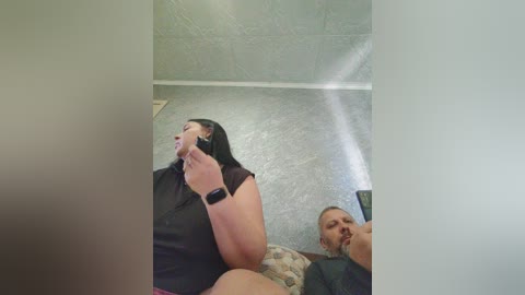 A candid video of a woman with long black hair, wearing a black shirt, talking on a phone, and a bearded man with a mustache in a dimly lit, textured gray room.
