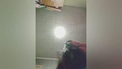 A video captures a young woman with long, dark hair, standing in a dimly lit room with a textured, light gray ceiling. She is looking up, with a hand in her hair. The room has a wooden ceiling beam and a small, bright light source.