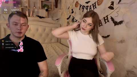 Video of a young woman with light skin and shoulder-length, straight, light brown hair, wearing a white top and black skirt, seated in a pink chair. A man with short, light brown hair, wearing a black shirt, stands next to her. The background features Halloween decorations.