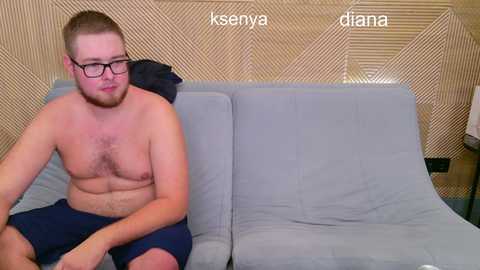 Video of a shirtless, muscular man with short brown hair, wearing glasses and dark shorts, sitting on a gray couch against a textured, beige wall. Text reads \"ksenia\" and \"diana.\