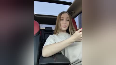 Video of a young Caucasian woman with long, straight blonde hair, fair skin, wearing a beige t-shirt, sitting in the driver's seat of a car with red and black interior.