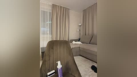 A video of a minimalist, beige-toned living room with a L-shaped sofa, sheer curtains, a white table, and a spray bottle on a carpeted floor.