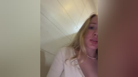 A video of a young blonde woman with fair skin and a slender physique, wearing a white cardigan, peering out from a small, dimly lit room with white paneled walls.