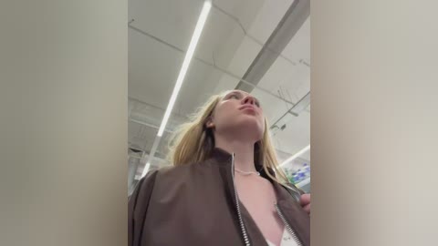 Video of a blonde woman with pale skin, wearing a brown jacket, standing in a brightly lit, modern office space with white walls and fluorescent lighting.